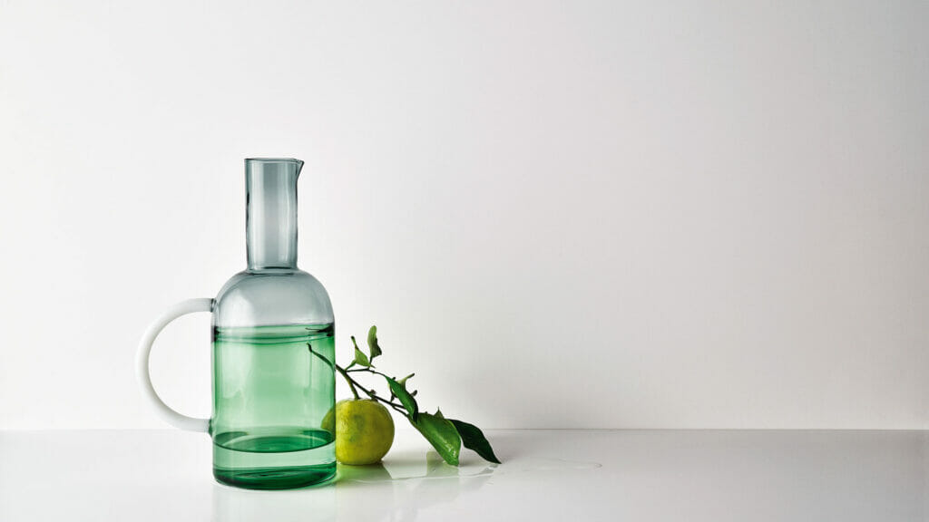 Stylish green glass water jug beside a lemon on a minimalist white background, showcasing modern elegance.