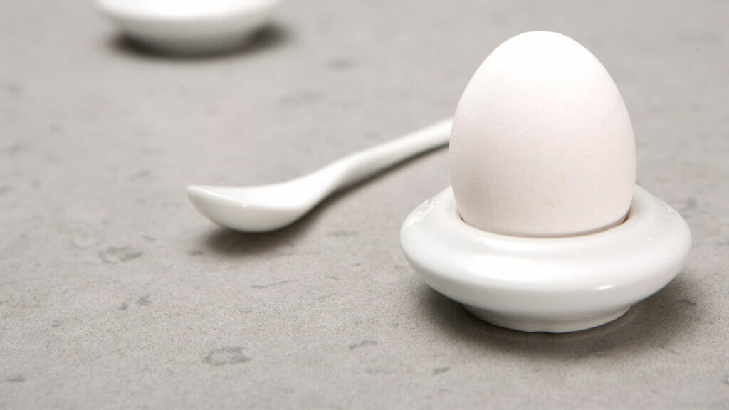 White egg in ceramic holder with a spoon on a light gray stone surface, emphasizing minimalist dining aesthetics.