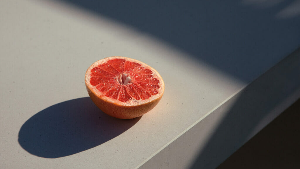 Freshly cut grapefruit half resting on a smooth concrete surface, showcasing vibrant pink flesh and realistic shadows.