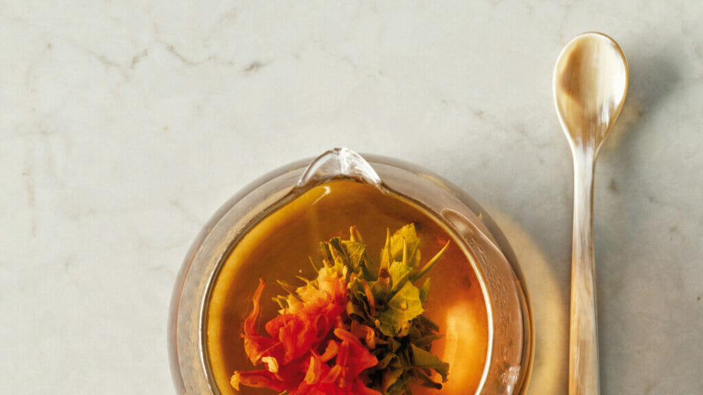 A clear bowl with a colorful floral tea infusion beside a silver spoon on a marble surface.