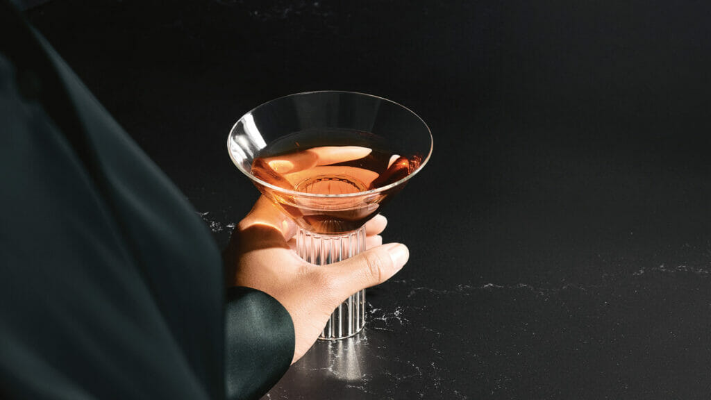 Person holding a stylish cocktail glass filled with pink drink on a dark, luxurious countertop surface.