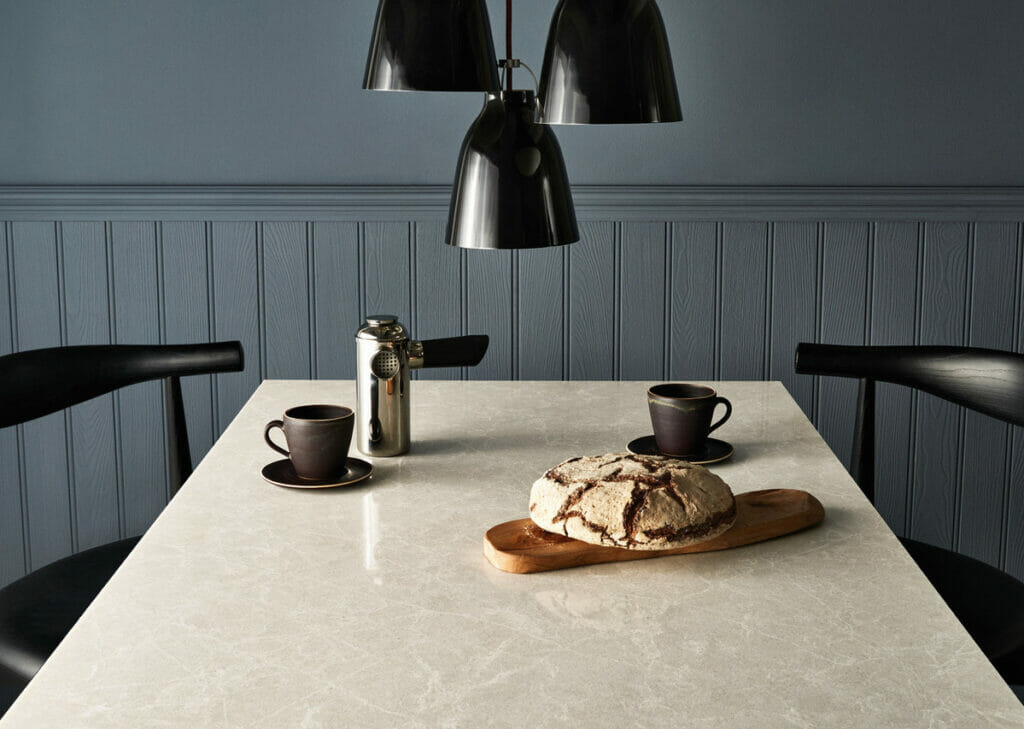 Elegant dining setup with coffee cups, a loaf of bread on a wooden board, and sleek black chairs against a light-colored table.