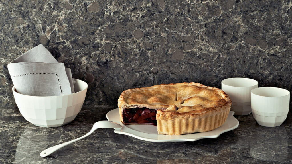 Delicious cherry pie on a white plate with bowls and napkins against a dark textured countertop.