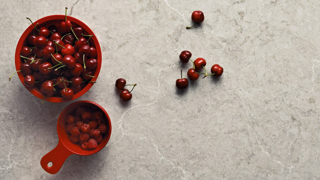 Red bowls filled with fresh cherries and raspberries on a light textured surface.