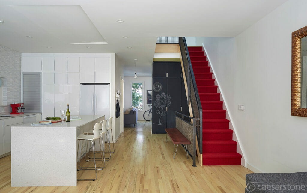Modern kitchen and living area with white cabinets, a red staircase, and wooden flooring, showcasing a bright, open space.