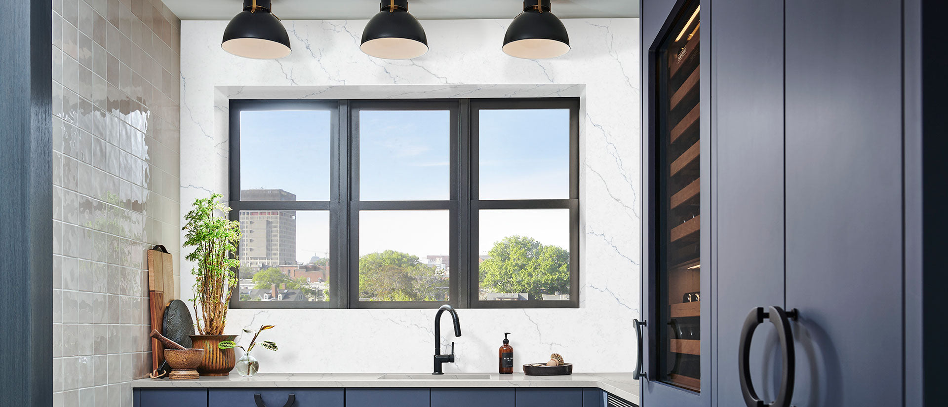 A contemporary kitchen featuring Calacatta Miraggio Cielo quartz countertops and a matching waterfall island, highlighting soft blue and grey clouded veining on a white base, paired with dark blue cabinetry and sleek gold hardware.
