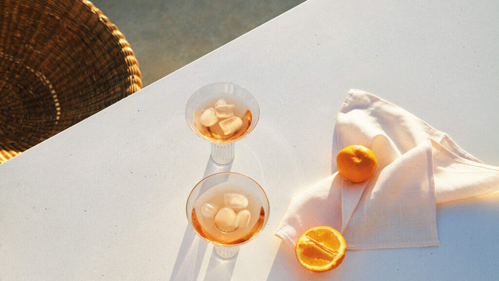 Two cocktails with ice sit on a white table, accompanied by oranges and a clean cloth, basking in warm sunlight.