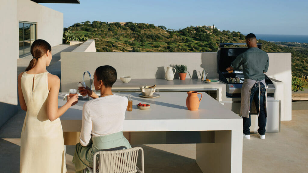Two women enjoying drinks at a modern outdoor kitchen with a man barbecuing, overlooking a scenic landscape.