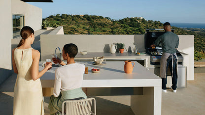 Two women enjoying drinks at a modern outdoor kitchen with a man barbecuing, overlooking a scenic landscape.