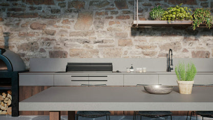 Modern kitchen featuring smooth concrete grey surfaces and stone wall, with plants and rustic oven in the background.
