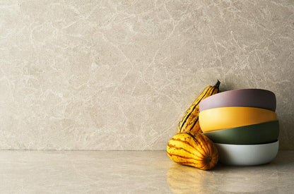 A stack of colorful bowls next to decorative squash on a cream-colored surface with elegant ashen streaks.