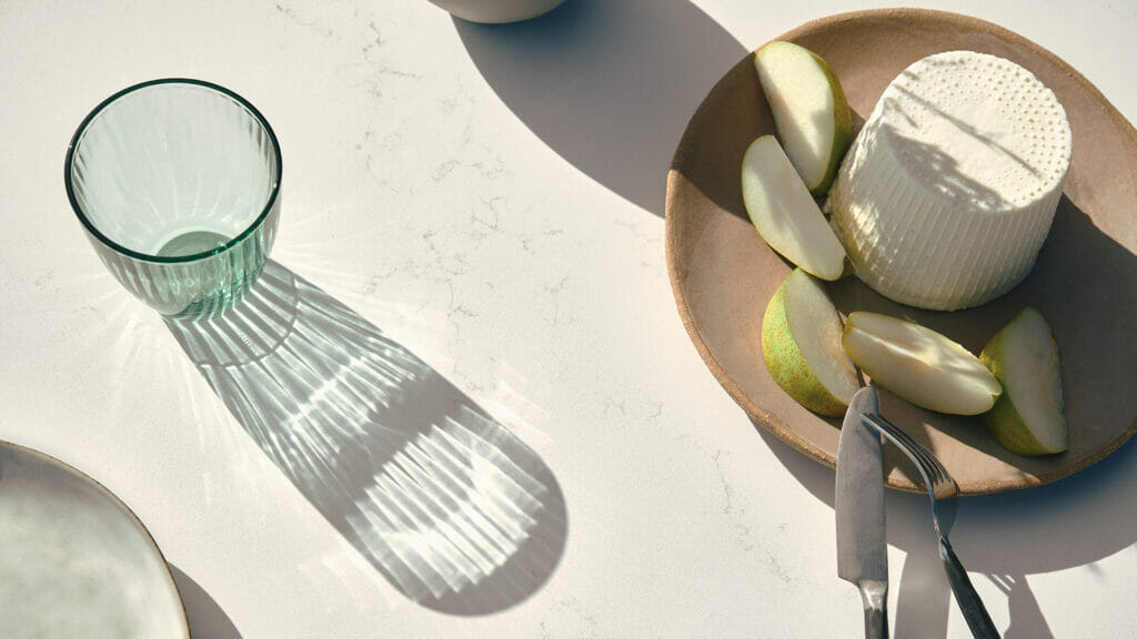 Elegant dining scene featuring a glass, pear slices, and cheese on a light background with play of shadows.